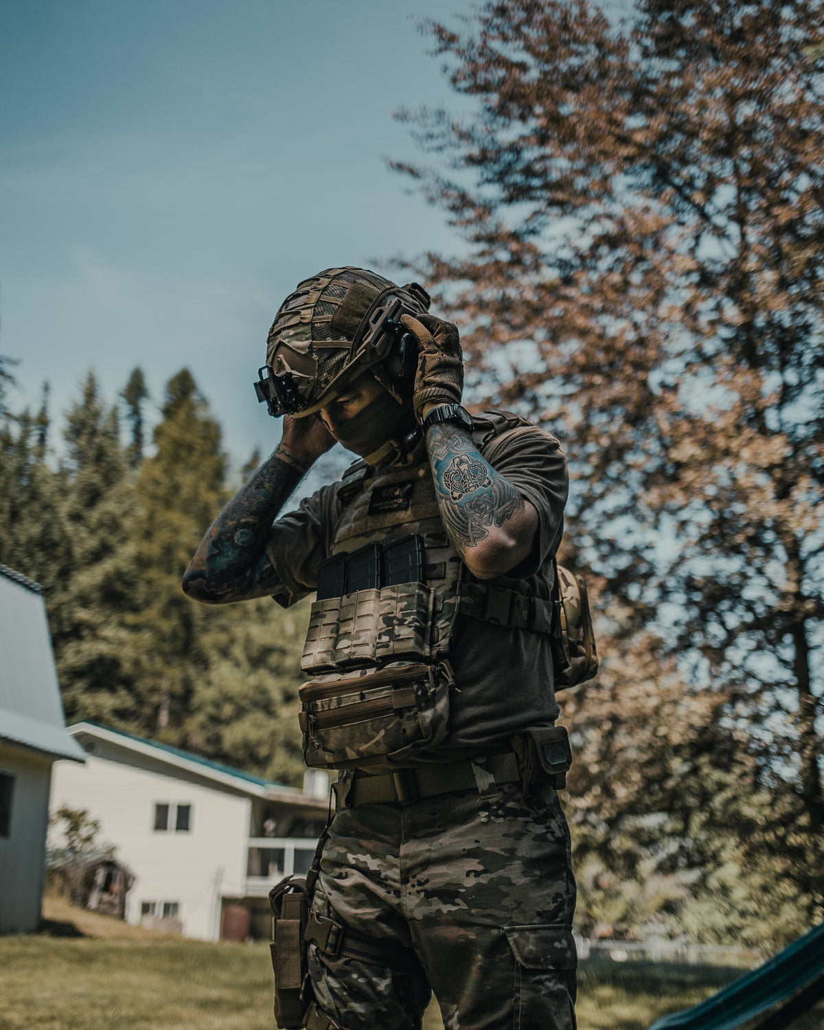 Person in military camouflage gear adjusting helmet outdoors with trees and building in background